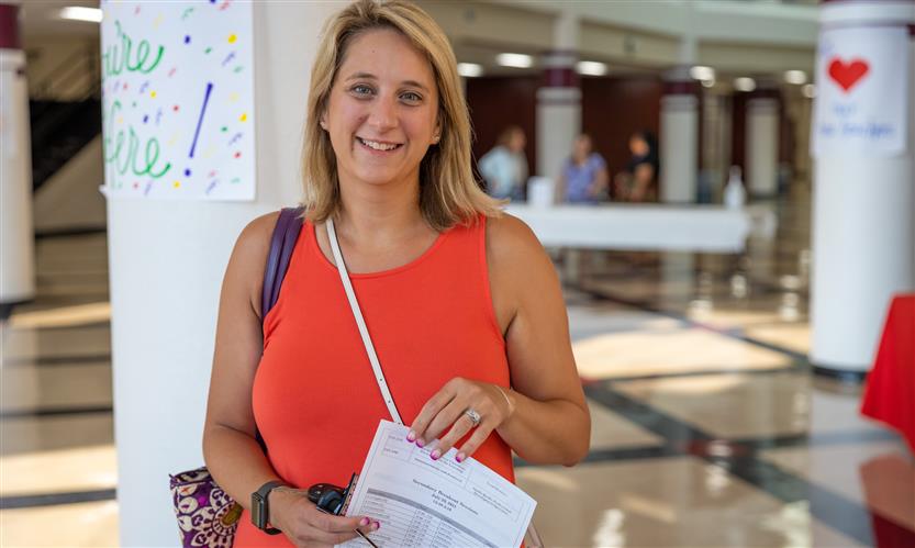 A woman in an orange shirt smiles while standing in a school hallway